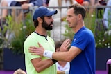Two male tennis players meet at the net after a match, the one on the right in discomfort, the other showing sympathy