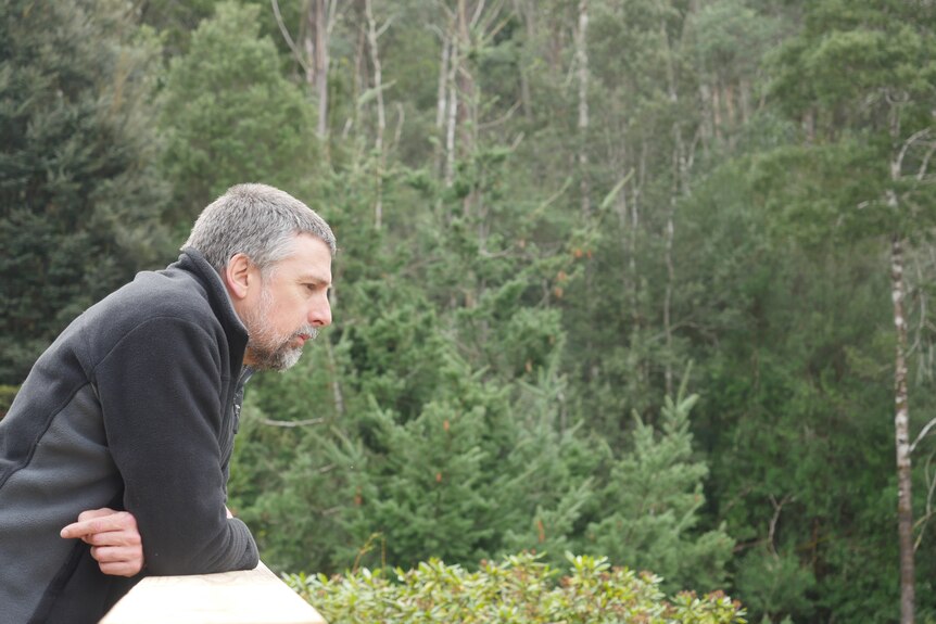 A man with close cropped grey-brown hair and beard leans against a railing and looks out over his bush property