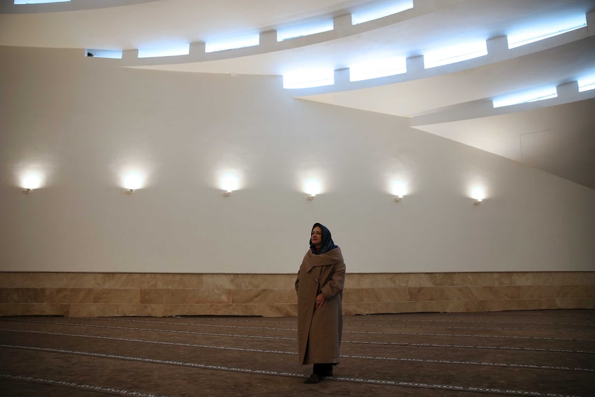 A veiled woman walks into a mosque's prayer hall featuring modern design of undulating waves of grey and concrete.