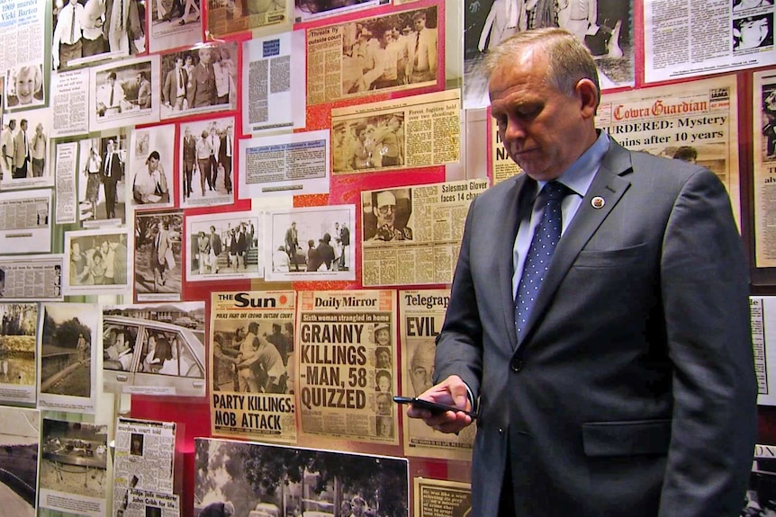 Detective Chief Inspector Grant Taylor stands in front of a wall of newspaper clippings while looking at his phone.