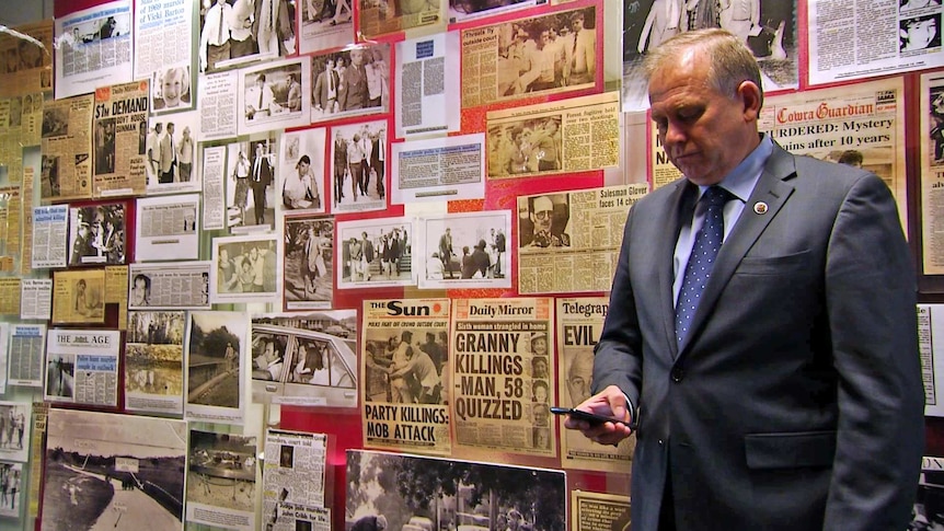 Detective Chief Inspector Grant Taylor stands in front of a wall of newspaper clippings while looking at his phone.