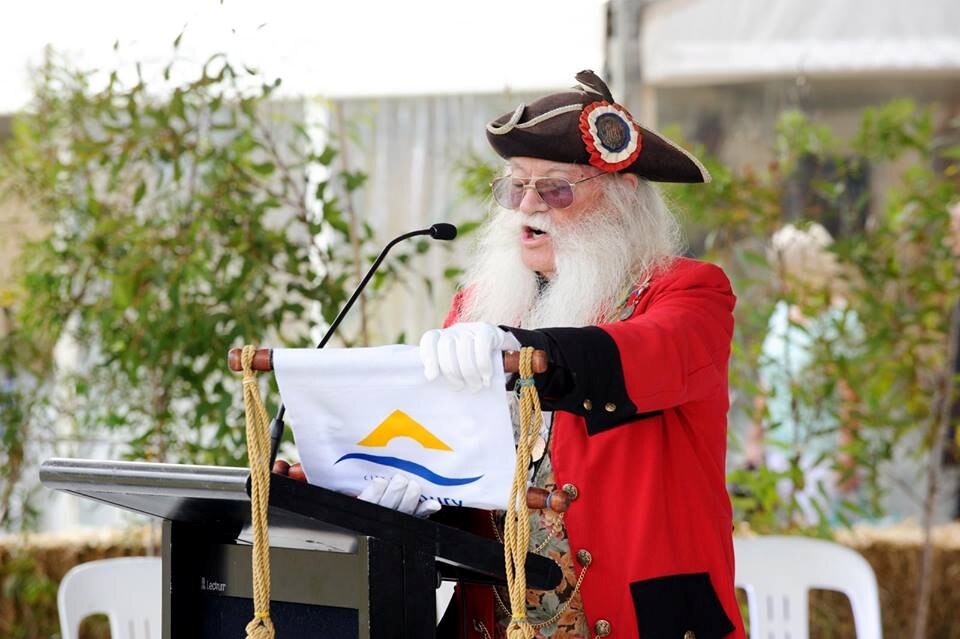 man in long white beard and sunglasses and black hat and red coat reading from a scroll