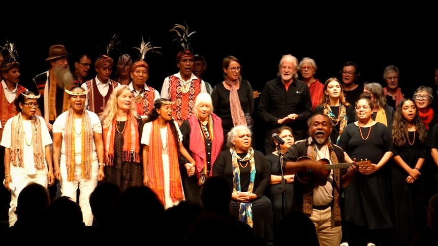 Two choirs sing together, with some members wearing traditional feather headdresses.