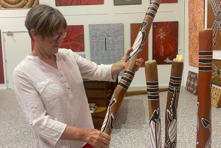 A woman standing in a room of paintings holding and looking at a didgeridoo