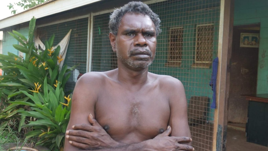 A photo of an Indigenous man crossing his arms in front of his house.