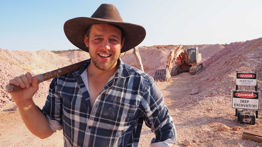 A man in a broad brown hat smiles as he holds a pick axe over his shoulder, standing in front of an excavator and dirt mounds