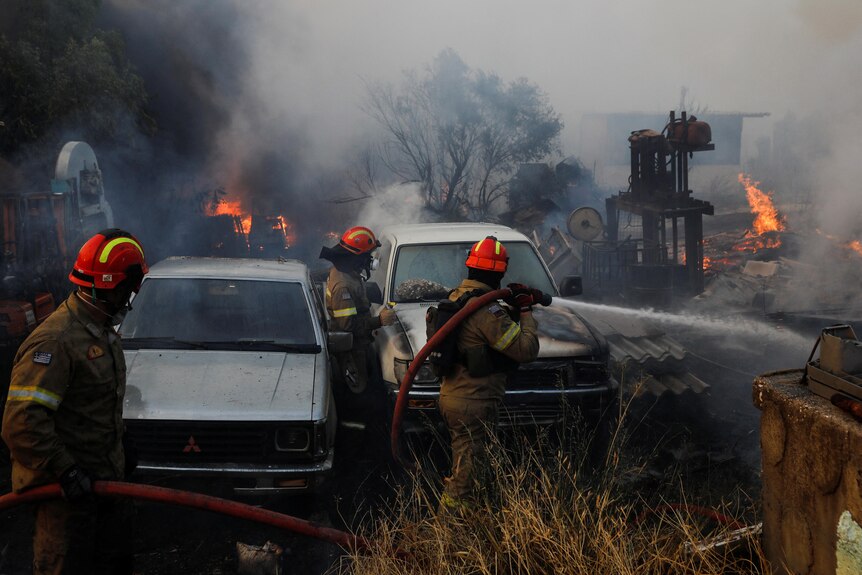 Trzech strażaków z wężem obok dwóch samochodów z dymem i płomieniami w tle. 