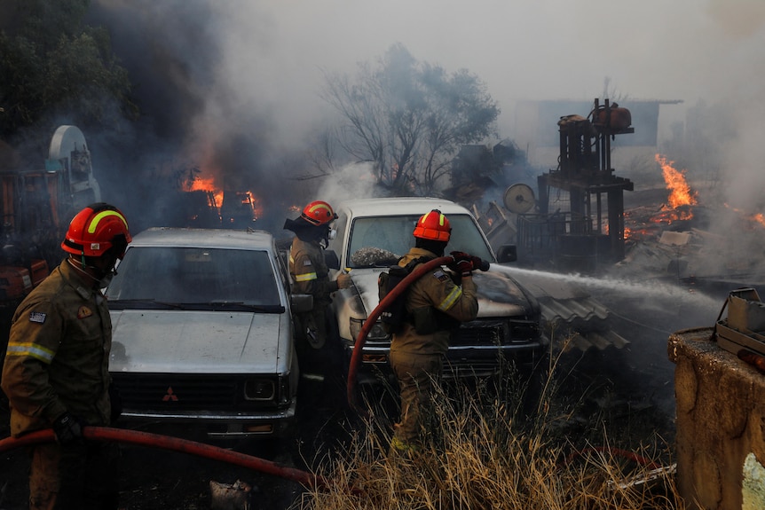 Three firefighters with a hose next to two cars with smoke and flames in the background. 