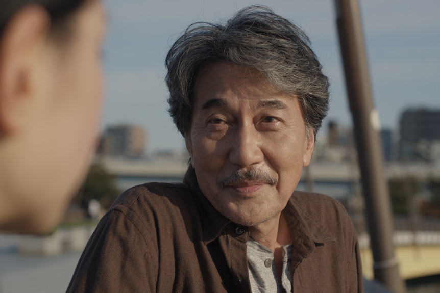 A film still of a close-up of Kōji Yakusho, a 68-year-old Japanese man, wearing a brown jacket, with a slight smile.