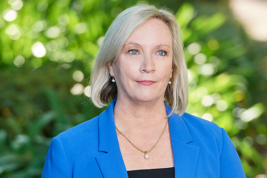 A middle-aged woman with fair, shoulder-length hair stands in front of some greenery.