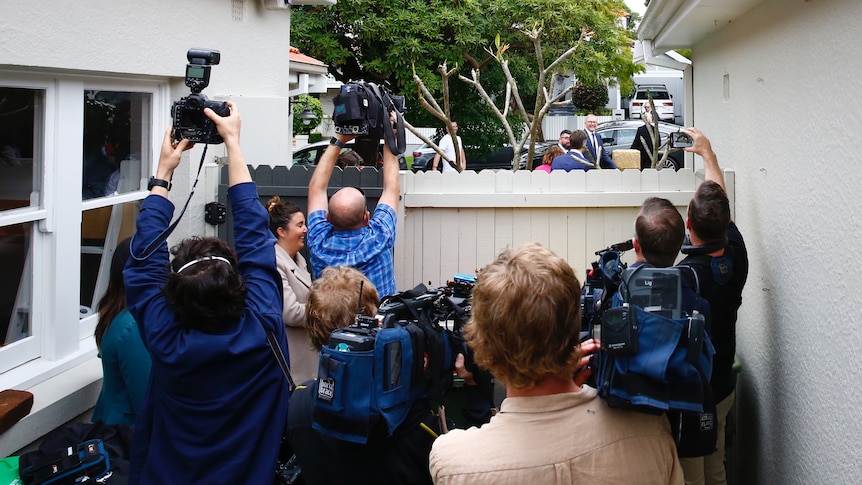 A group of people with cameras stand behind a fence as Anthony Albanese and staff walk past on the other side. 