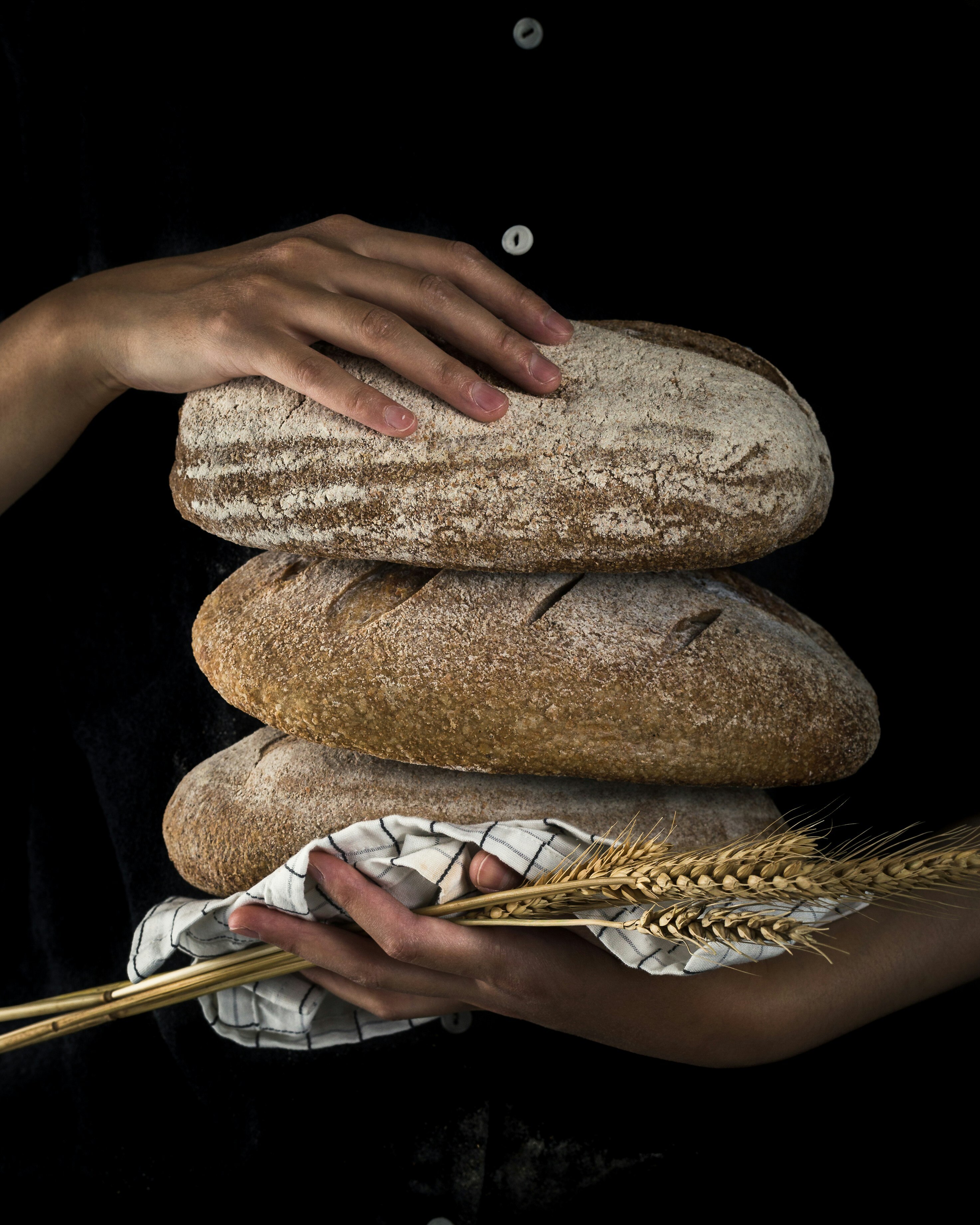 A pair of hands holds several stems of wheat and three loaves of bread, stacked.