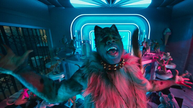 A CGI cat with man's face sings with arm outstretched in a blue a pink neon lit club space, small cats dance behind him.