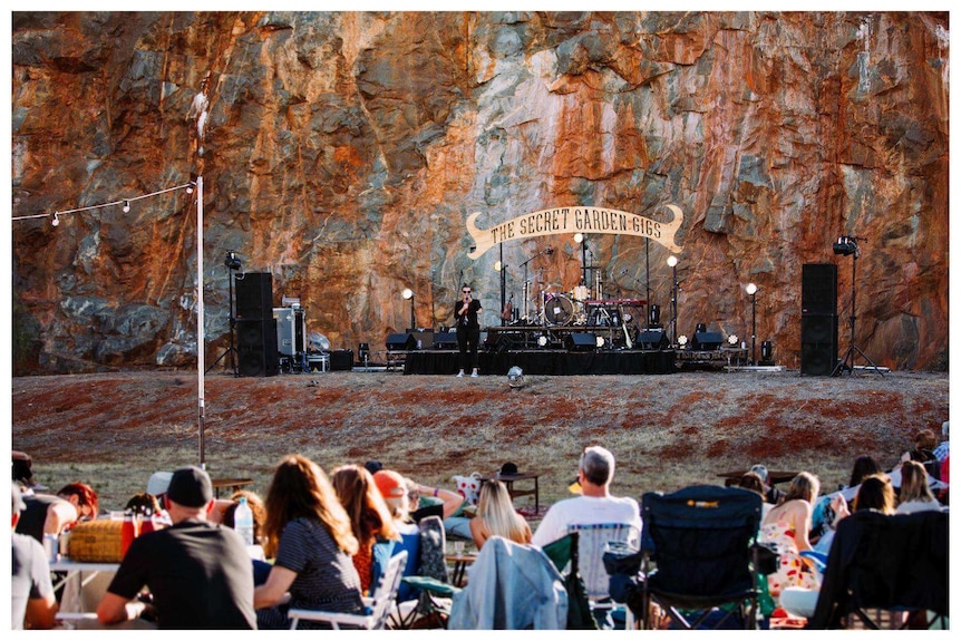 A crowd watches a person standing on a stage set at the foot of huge wall of sheer, red rock.