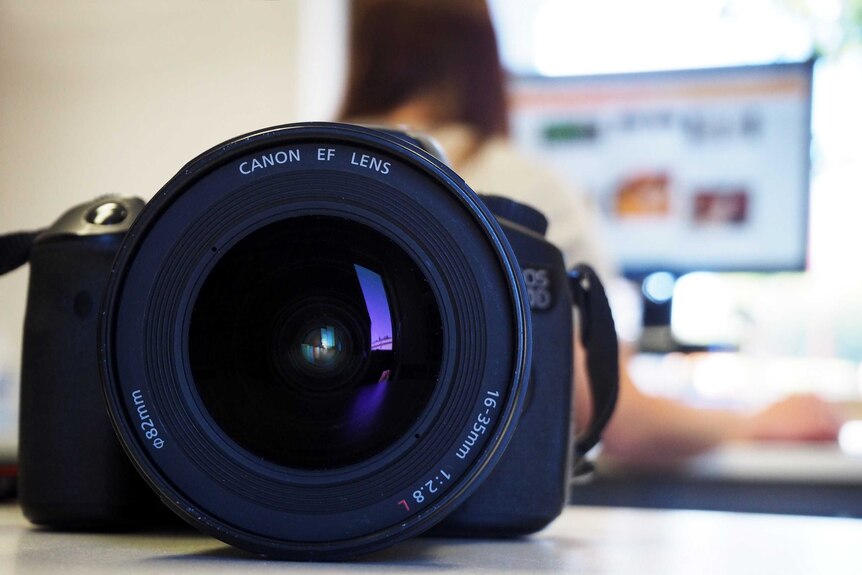 Close up photo of a camera, looking directly at the lens, with a person working at a computer out of focus in the background.