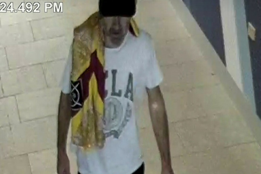 A man is captured on CCTV walking into a hospital, towel on his shoulder, wears a cap, white tee with something written on it.