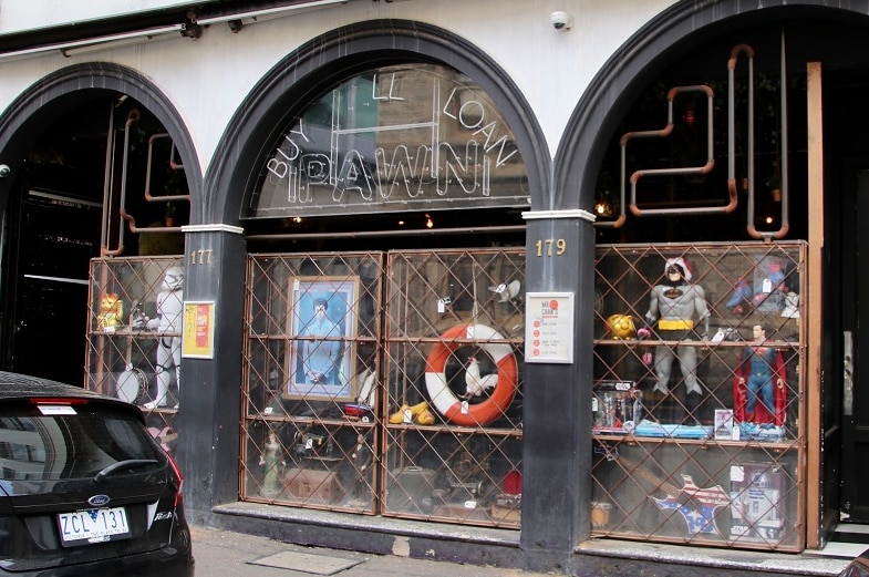 The outside of a nightclub is set up to look like a pawn shop with second hand paintings and action figures in the window.