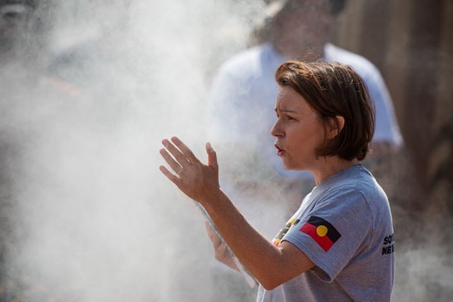 Woman in t-shirt with Aboriginal flag stands at a smoking ceremony