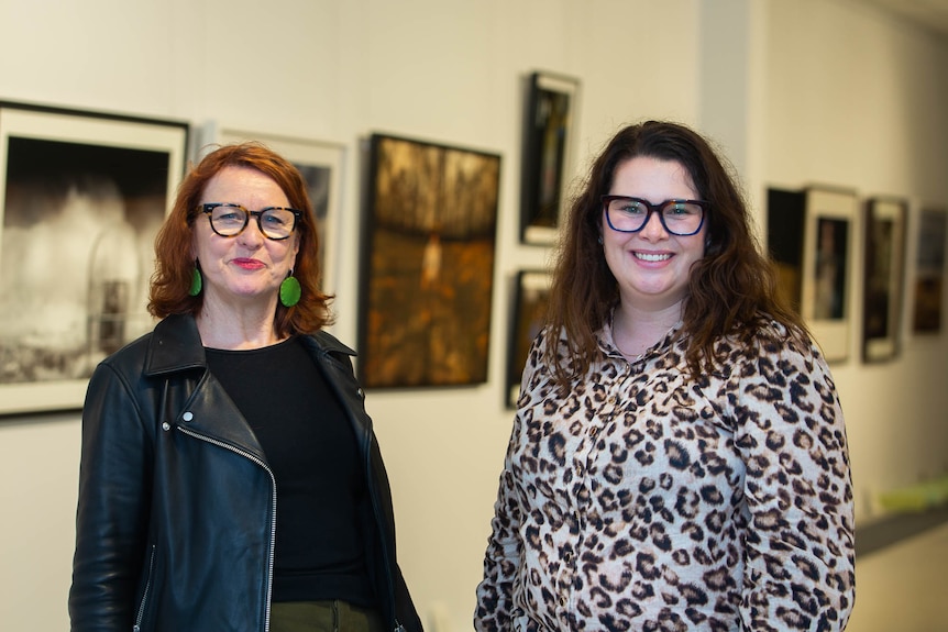 Dos mujeres sonriendo a la cámara con una pared de galería de fotografías detrás