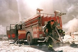 A firefighter is seen wearing black with a hose on his shoulder, with a truck in the background and dust in the air.