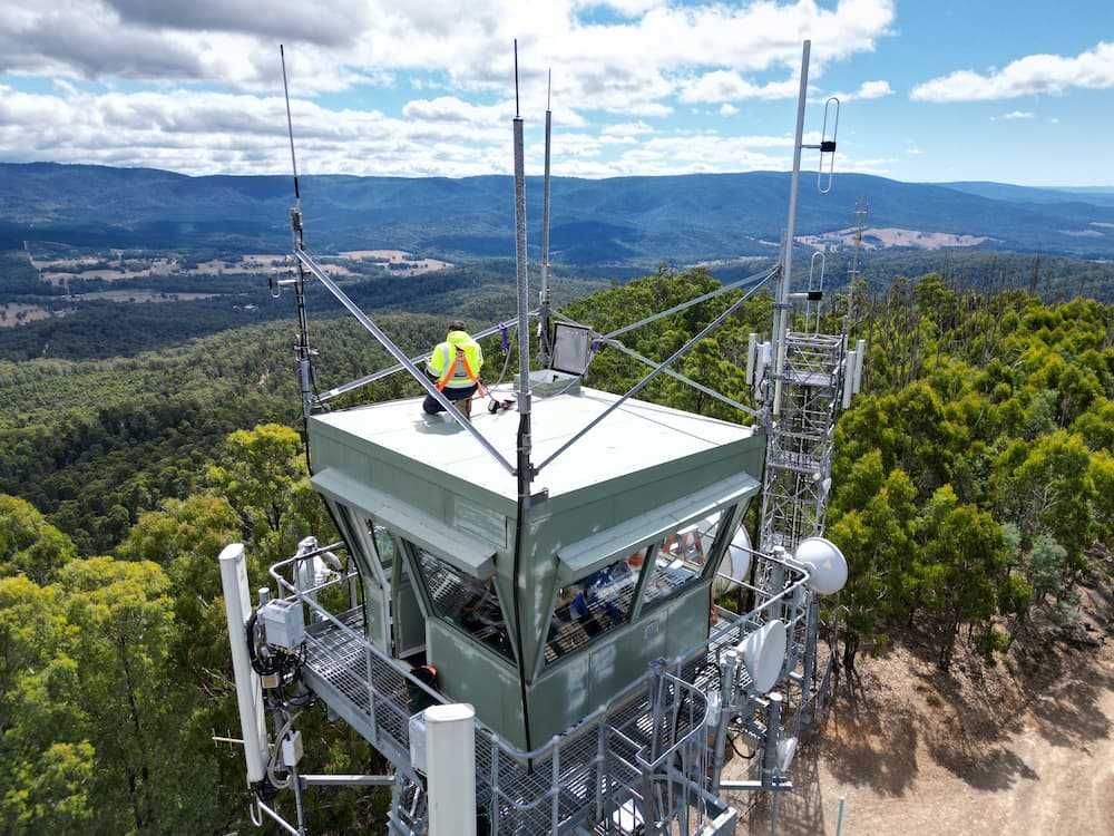 A fire lookout surrounded by bush with a man in a fluro vest couching at the top of the lookout,