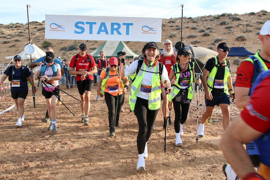 A group of runners, some in high vis vests and hiking poles walk towards the camera, away from a 'start' banner and a sand dune.