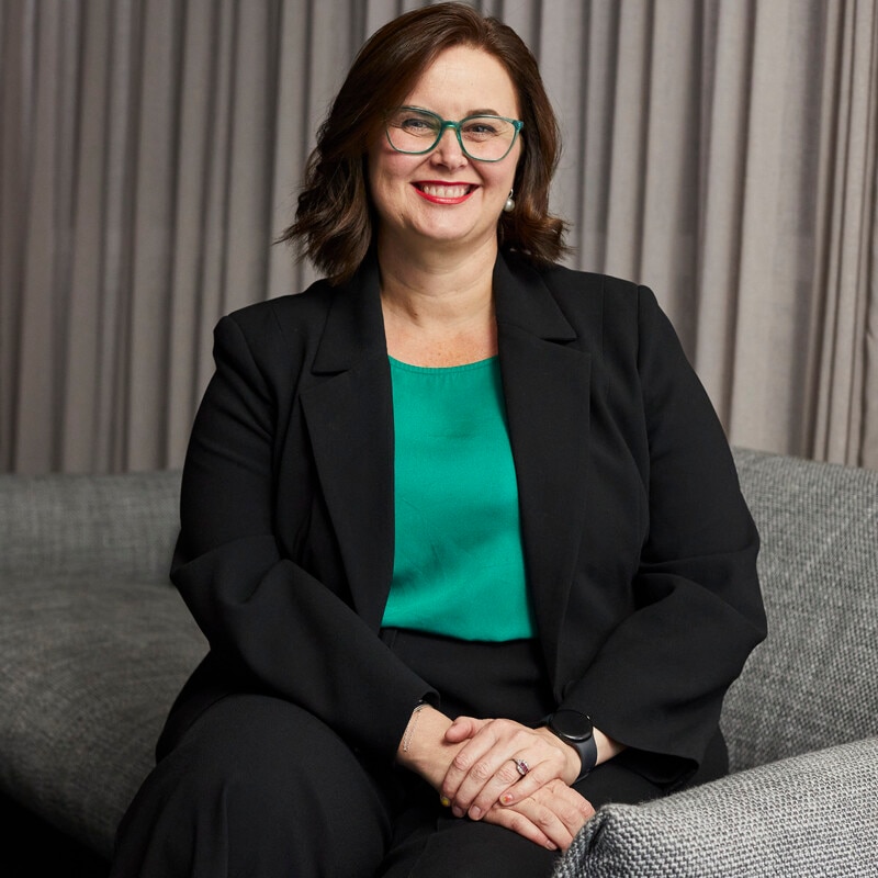 Cara Varian con gafas verdes, top verde, blazer negro, bob moreno, sonriendo.
