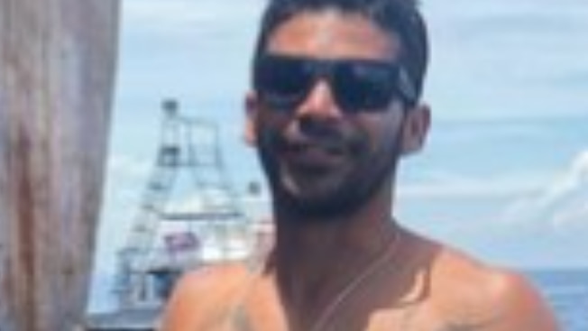 A young man, shirtless and tattooed, wearing dark sunglasses and smiling as he stands with his back to the ocean.