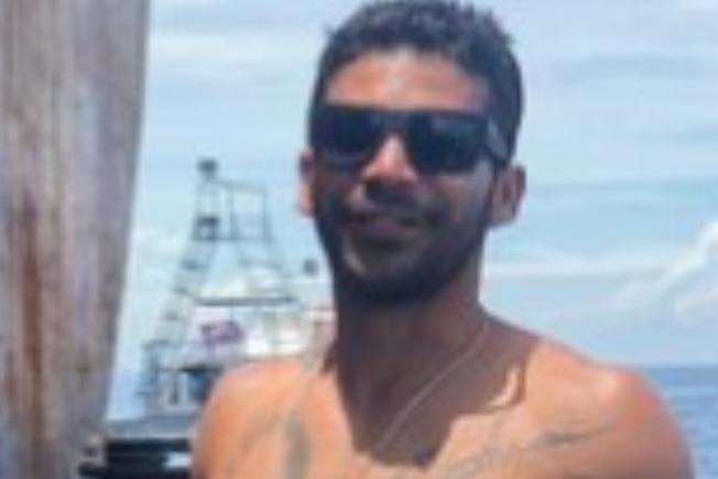 A young man, shirtless and tattooed, wearing dark sunglasses and smiling as he stands with his back to the ocean.