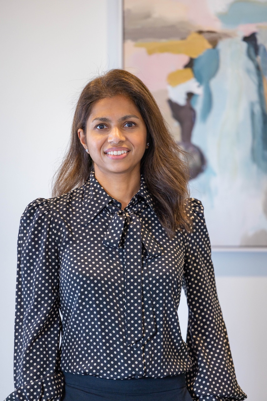 Uma mulher de cabelos castanhos e camisa de bolinhas em frente a uma pintura em tons pastéis.