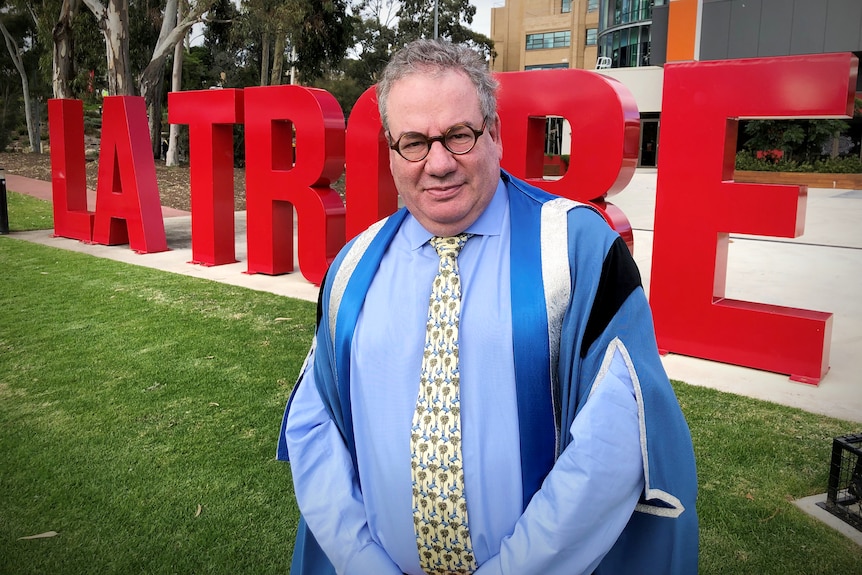 A grey-haired man stands in graduation robes in front of a sign that reads la trobe
