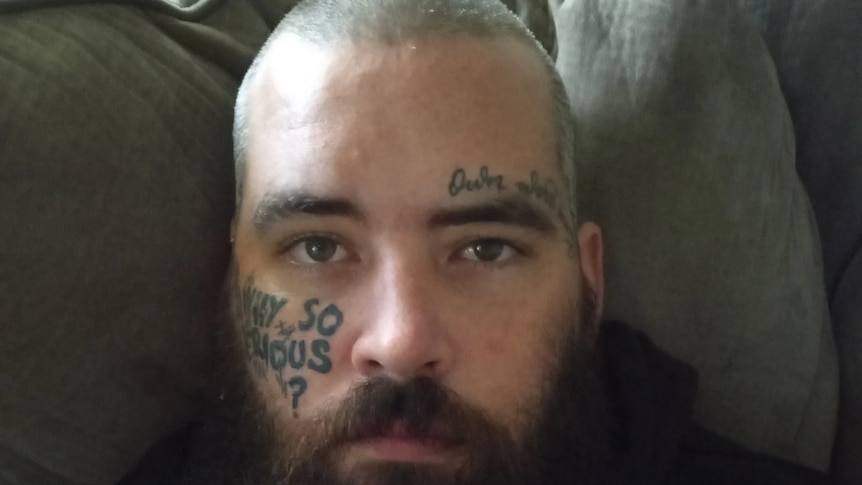 A bald, bearded white man in his late 20s with tattoos on his face.