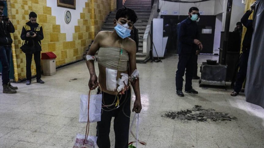 A badly injured man stands holding containers of blood.