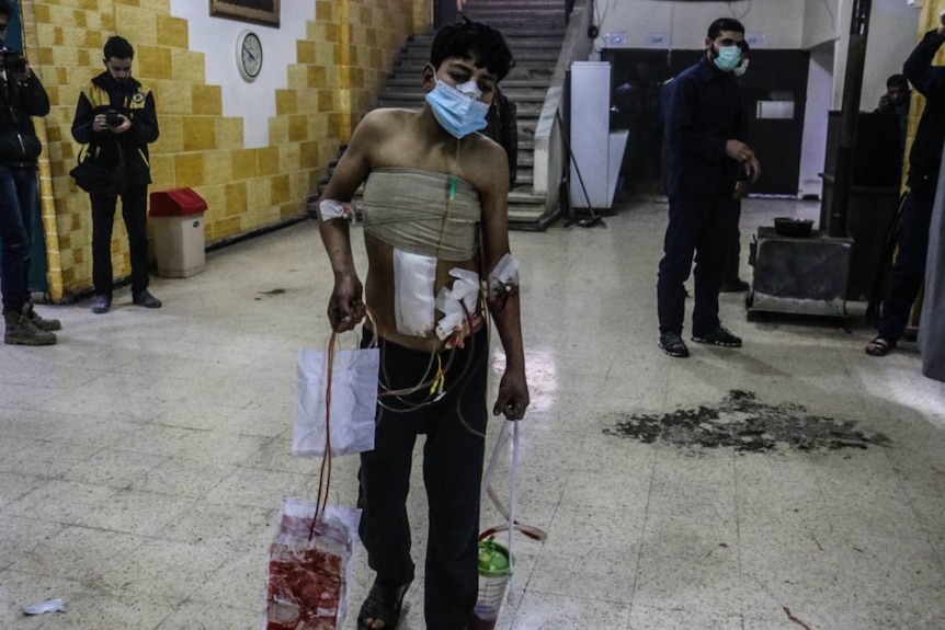 A badly injured man stands holding containers of blood.