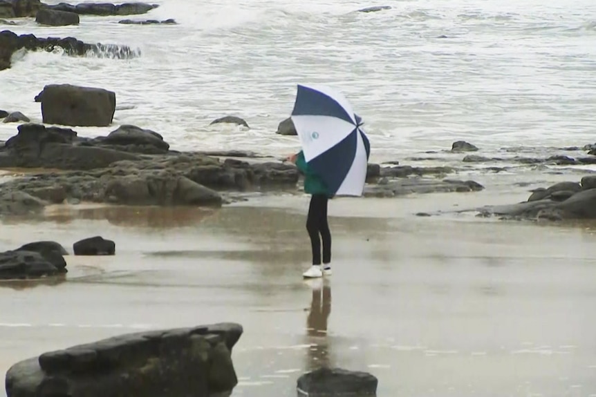A person stands on the beach with a blue and white umbrella.