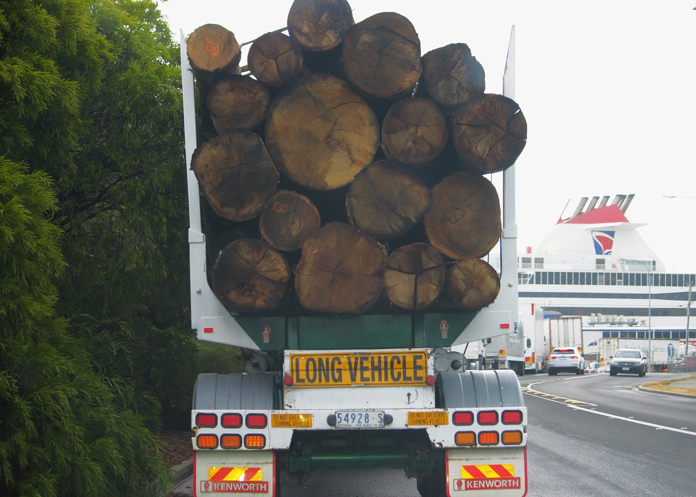 A semi-trailer loaded with logs in a traffic lane approaching a port. The large Spirit of Tasmania ship is in front of it.
