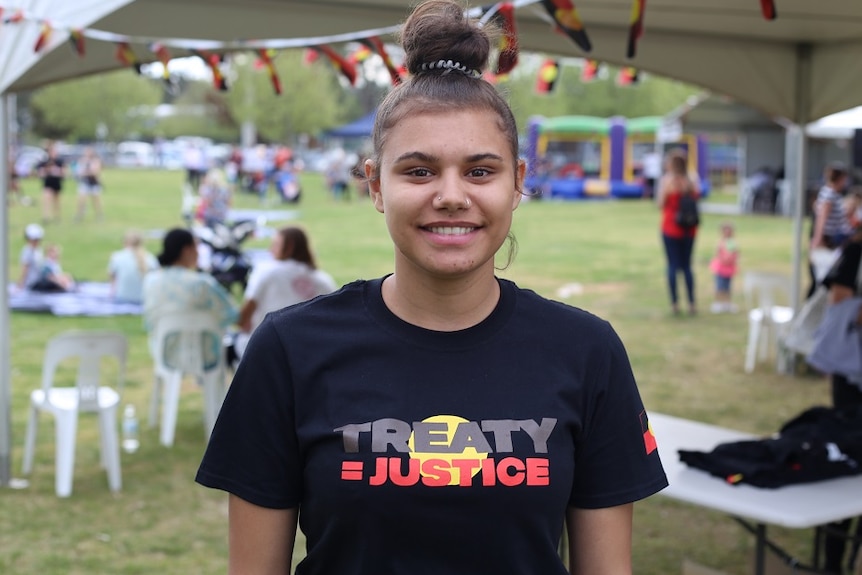 Nikita smiles as she stands in front of a tent adorned with Aboriginal flags on a grassed area.
