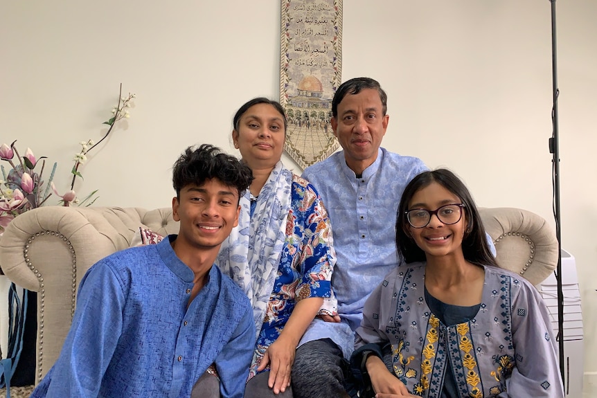 Mashreka, her husband and two kids smile at the camera, on the wall is a small hanging featuring an Islamic shrine.