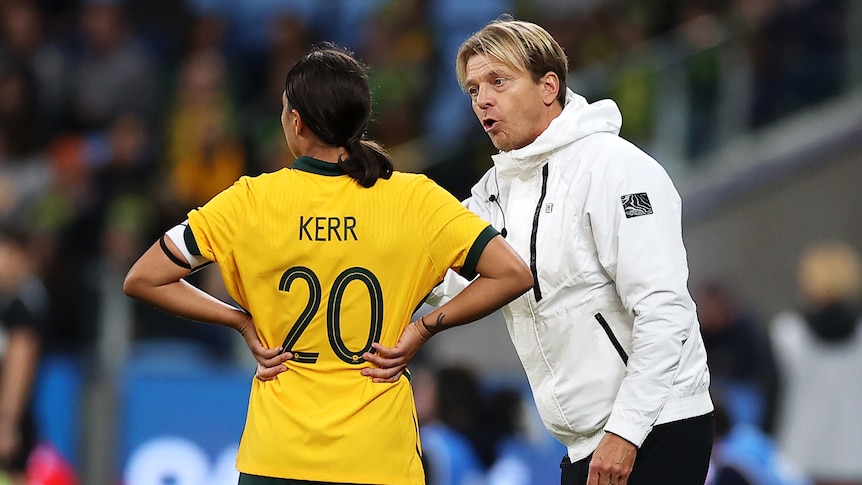 A man in a white jacket speaks to a woman soccer player wearing yellow and green