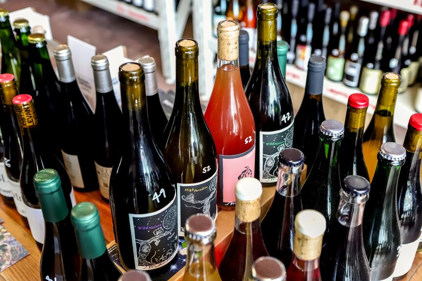 Row of brightly coloured bottles of wine sit on bench inside wine store with racks of wine bottles in background