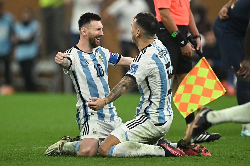 Two soccer players hold their arms widely as they prepare to hug while kneeling on the pitch.