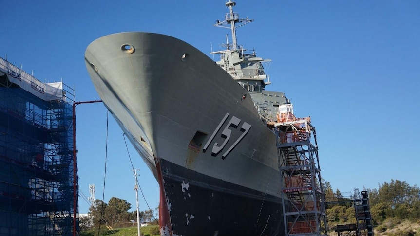 A large grey warship sits on supports in dry dock