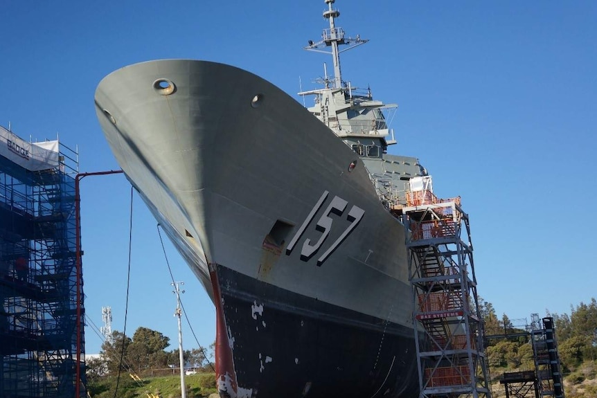 A large grey warship sits on supports in dry dock