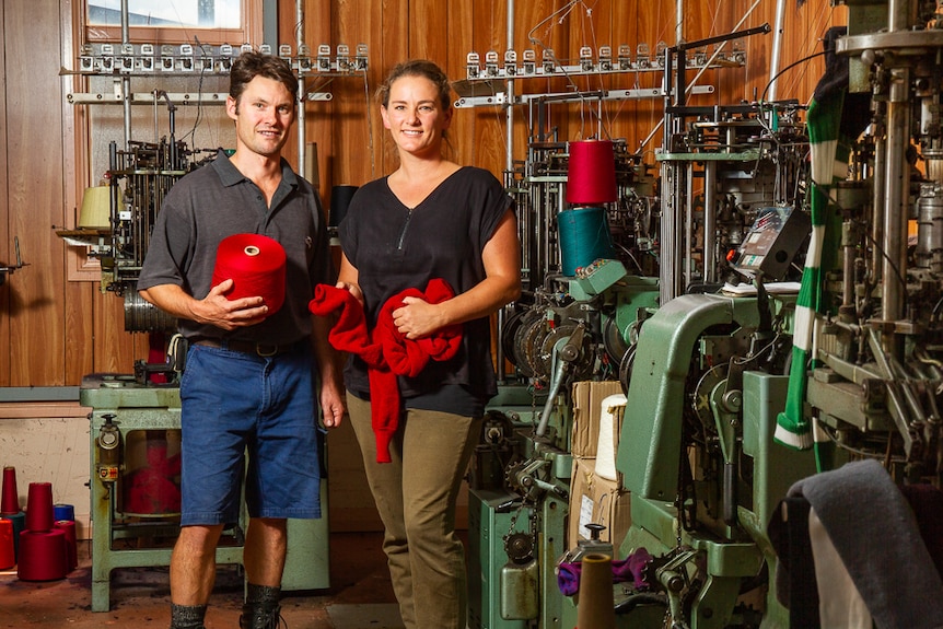 A man and woman holding wool and socks while standing next to knitting machinery.