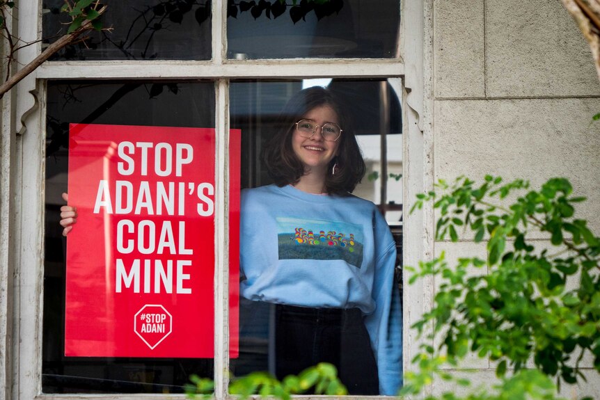 Jean Hinchliffe inside her house seen through a window, she is holding a sign calling for the halting of the Adani development.