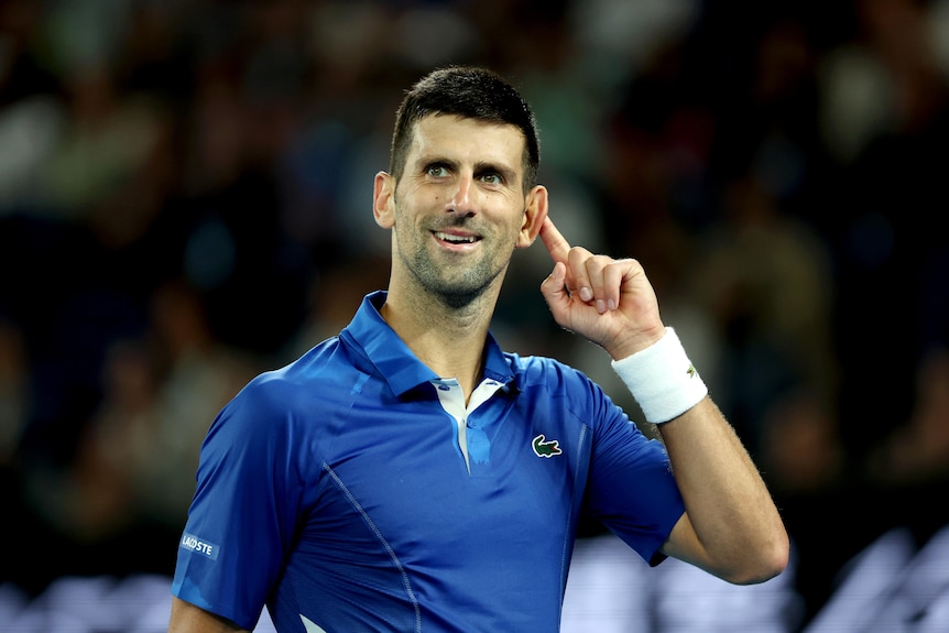 A male tennis player, wearing blue, holds his ear, gesturing to spectators to be loud