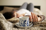 A woman lies on a bed with a tea cup and saucer in the foreground.