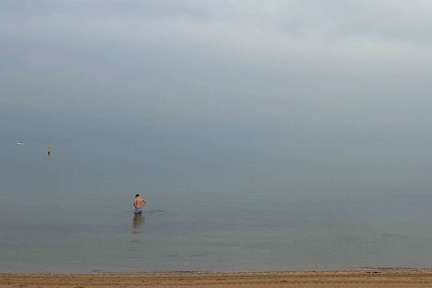 A male swimmer walks in the water in front of a bank of sea fog.