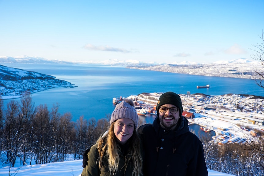 A couple stand with arms around each other smiling in front of a lake surrounded by snow.