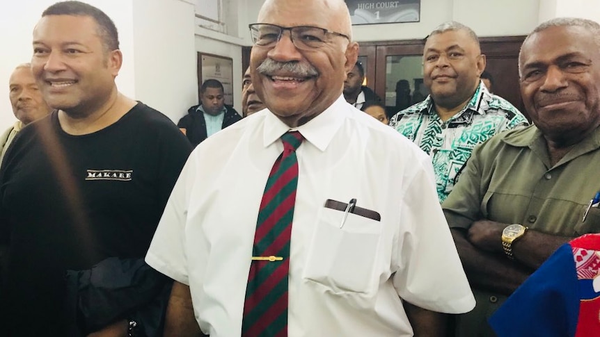 Sitiveni Rabuka smiles at the camera as he walks with a group of people out of court.